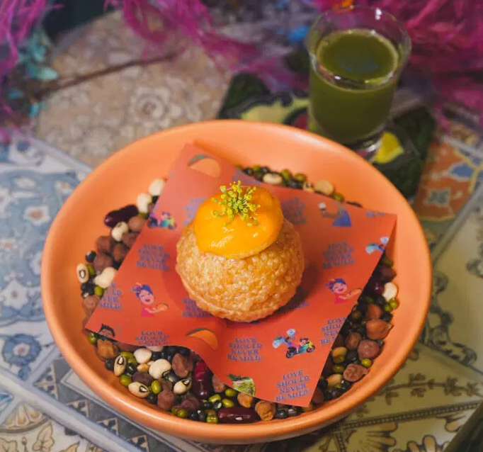 A colorful gourmet Indian dessert served on a printed sheet, placed over assorted lentils and beans in a terracotta bowl, accompanied by a shot of green herbal drink in the background.